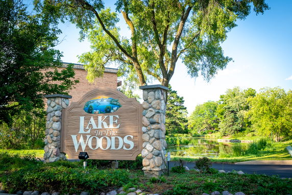 Sign reading 'Lake in the Woods' with trees and a small body of water in the background.