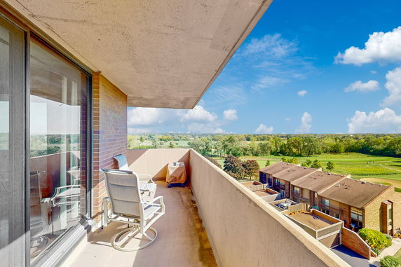 A balcony view from a high-rise building overlooking a suburban area with adjacent townhouses and greenery in the distance.