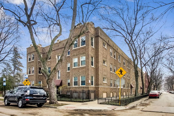 Street view of a multi-story brick apartment building with numerous windows and a metal fence along the sidewalk.