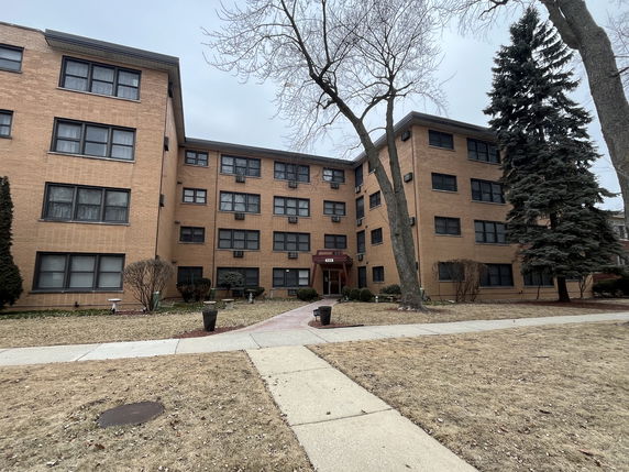 Front view of a multi-story apartment building with trees and shrubs in front.