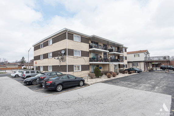 Front view of a multi-story apartment building with balconies and adjacent parking area.