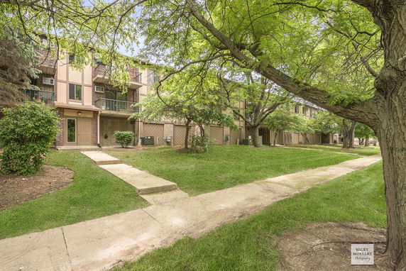 Front view of a residential multi-story building with balconies, surrounded by trees and greenery.