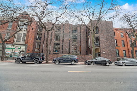 Front view of a multi-story brick building with balconies and parked cars in front.