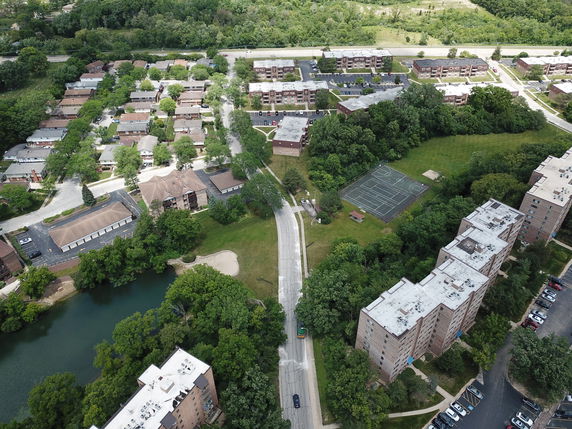 Aerial panoramic view of a residential area with multiple buildings, a road, and a sports court amidst greenery.