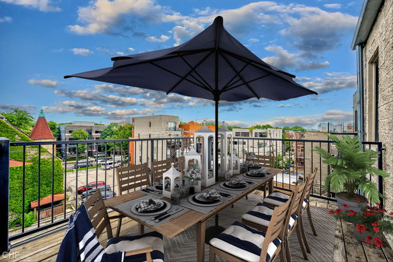 Rooftop dining area with a long table and umbrella, offering a view of surrounding buildings and a partly cloudy sky.