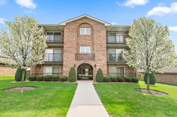 Front view of a three-story brick apartment building with balconies and landscaped surroundings.