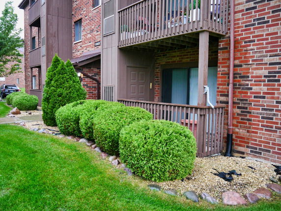 Front view of a building with brick and wood facade, featuring balconies and landscaped bushes.