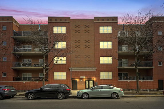 Front view of a multi-story apartment building with balconies and cars parked in front.
