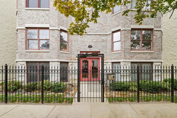 Front view of a multi-story brick building with red-framed windows and a wrought iron fence.