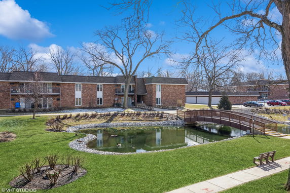 Front view of a multi-story brick apartment building with a small bridge over a pond in the foreground.
