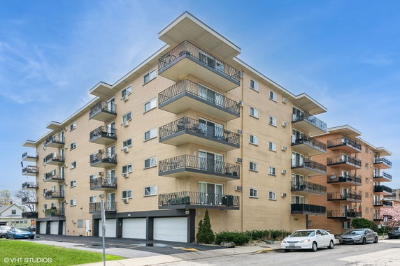 Front view of a multi-story apartment building with balconies and parking at the ground level.