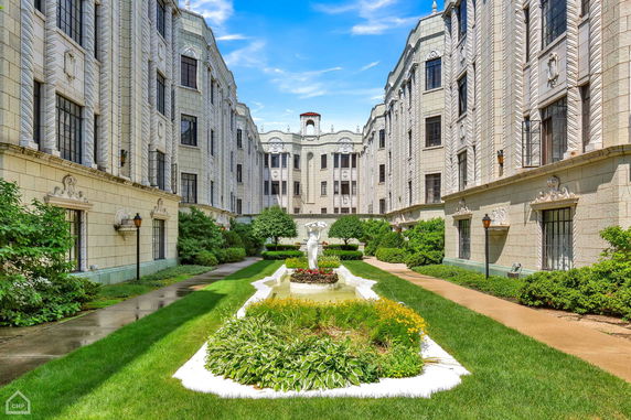 Front view of a multi-story building with ornate architecture and a landscaped garden with a statue in the courtyard.