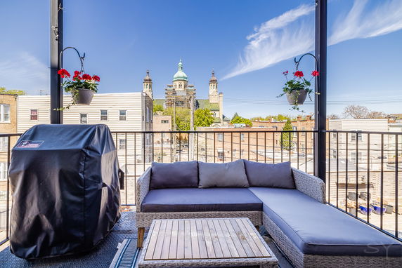 View from a balcony with outdoor seating and a grill, overlooking residential buildings and a church in the distance.