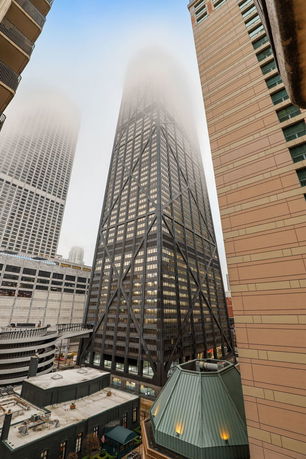 Tall skyscraper with distinctive exterior frame amidst other buildings, partially obscured by fog.