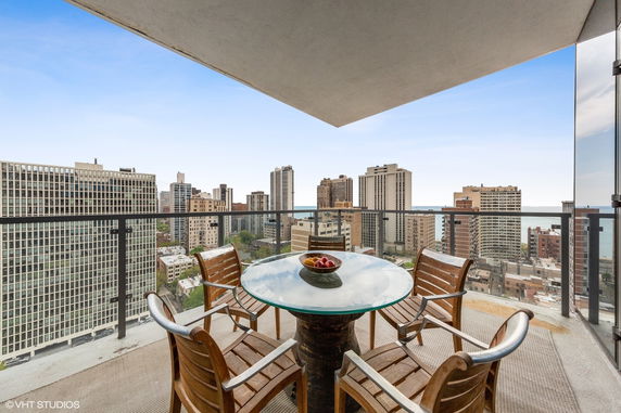 Balcony with a round glass table and chairs overlooking a cityscape with tall buildings.