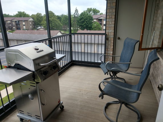 Balcony with a grill and chairs overlooking surrounding buildings and trees.