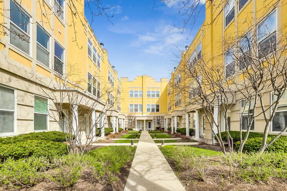 Yellow apartment buildings with multiple windows and a central walkway surrounded by landscaping.