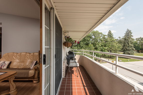 Balcony with railings, outdoor seating, and hanging plants, adjacent to an interior living area, overlooking a street and trees.