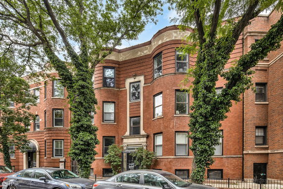 Front view of a brick apartment building with multiple windows and ivy-covered trees.