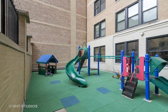 Indoor play area with playground equipment including a slide and playhouse set against a brick building wall.