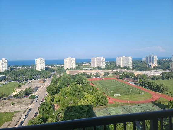 Panoramic view of a cityscape with buildings, a track and field, and a distant body of water.