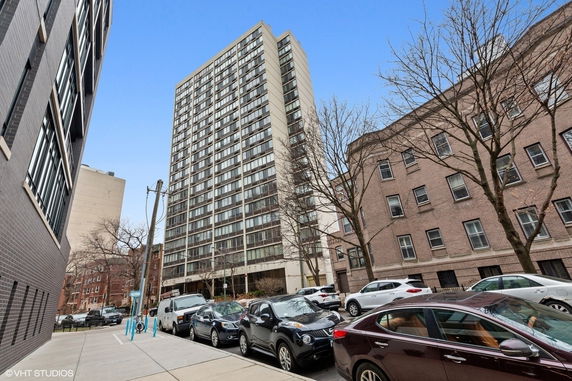 Front view of a high-rise residential building with multiple windows and a street with parked cars.