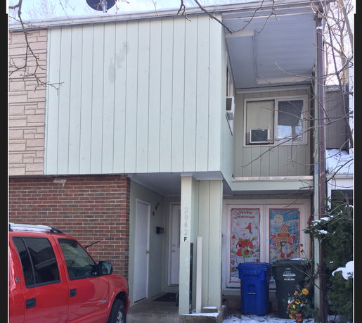 Front view of a two-story house with a brick and wood exterior, featuring a covered entrance and a second-floor window with an air conditioner.