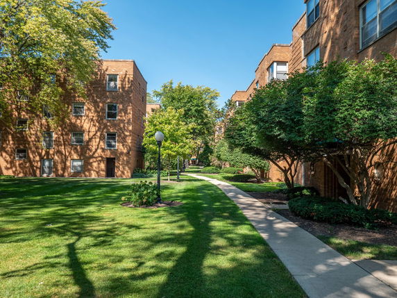 View of a brick apartment building with multiple windows and a walkway surrounded by greenery.