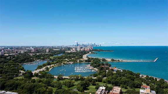 Panoramic view of a city skyline with lakes, green areas, and an expanse of water under a clear blue sky.