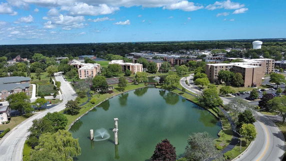 Aerial panoramic view of buildings surrounding a pond with a fountain in a landscaped area.