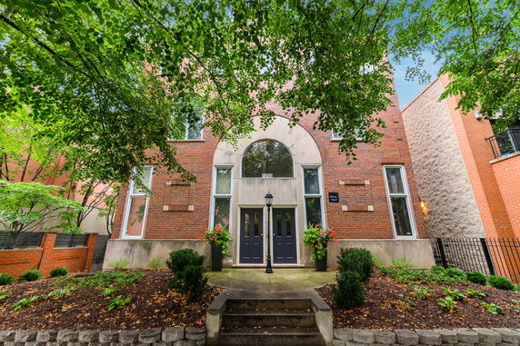Front view of a brick building with a central arched window and double doors, surrounded by greenery.