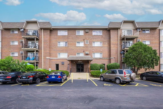 Front view of a multi-story brick apartment building with balconies and a parking area in front.