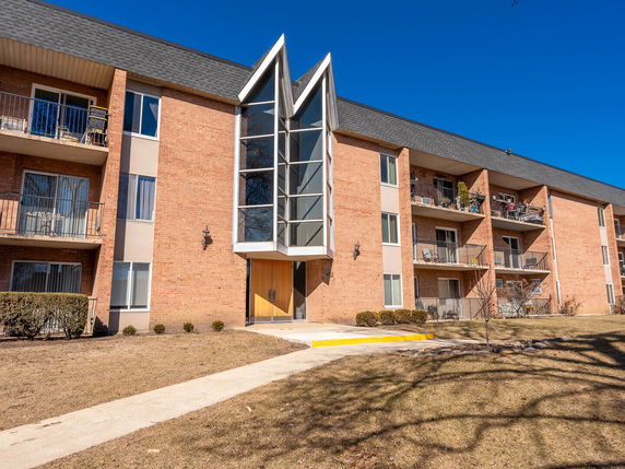Front view of a multi-story apartment building with balconies and a central entrance featuring large vertical windows.