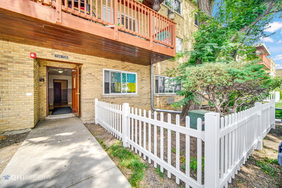 Front view of a multi-story brick apartment building with a white fence and wooden balcony.