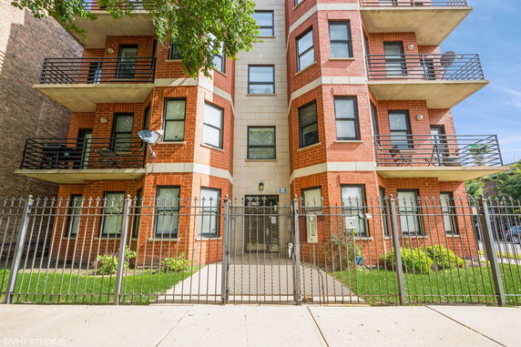 Front view of a multi-story brick building with balconies and a fenced entrance.