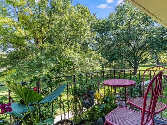 View from a balcony with red metal table and chairs, overlooking lush green trees and grass.