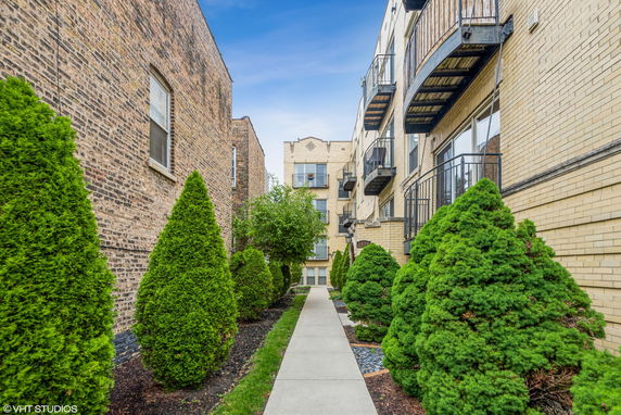 View of a multi-story brick apartment building with balconies and a landscaped walkway.