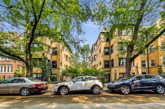 Front view of a multi-story apartment building with tree-lined street and parked cars.