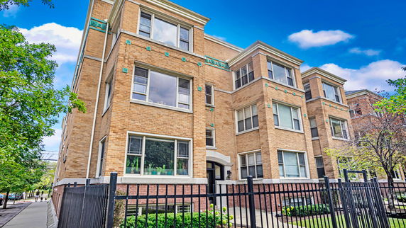 Front view of a multi-story brick apartment building with large windows and a fenced entrance.
