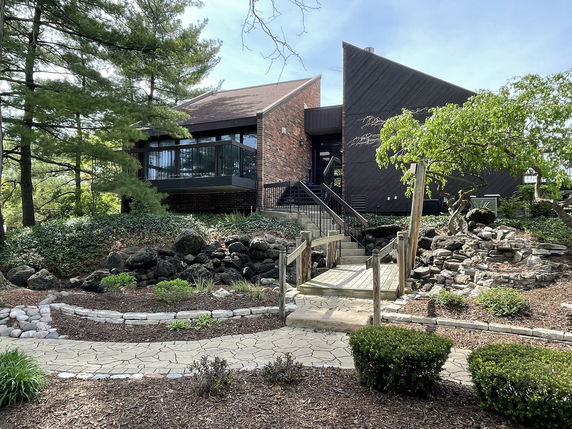 Front view of a modern house with a unique angular design and large windows, set amidst landscaped greenery.
