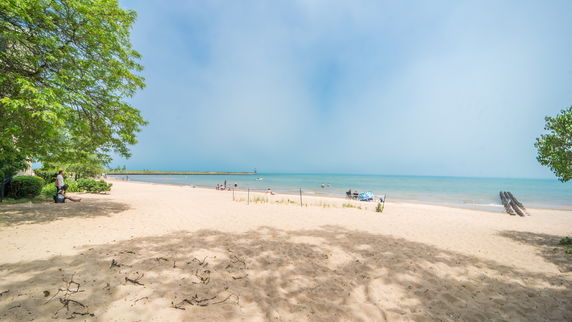 View of a sandy beach with people relaxing and swimming, bordered by calm blue water and distant trees.