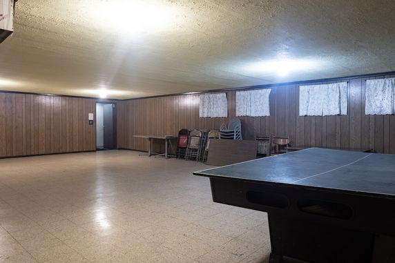Indoor recreational room with wood panel walls and tiled floor, containing a ping-pong table and stacked chairs.