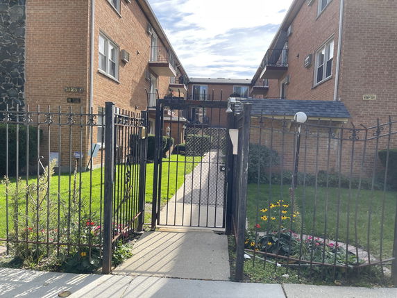 Front view of a multi-story brick building with balconies, fenced pathway, and small garden area.
