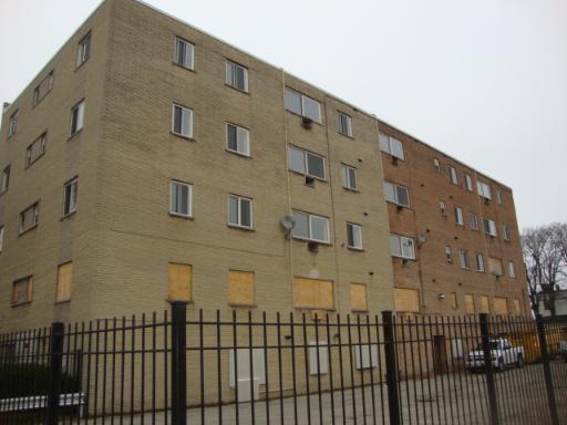 Front view of a multi-story brick building with boarded-up windows and a fenced perimeter.