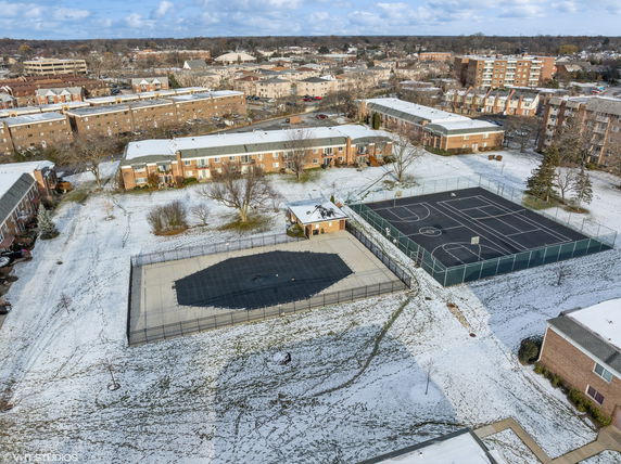 Aerial view of an outdoor play area with a basketball court and adjacent open space in a snowy residential neighborhood.