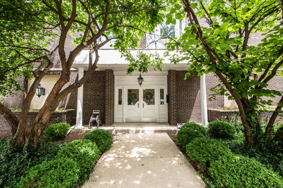 Front view of a building entrance with double doors and brick walls, surrounded by greenery.