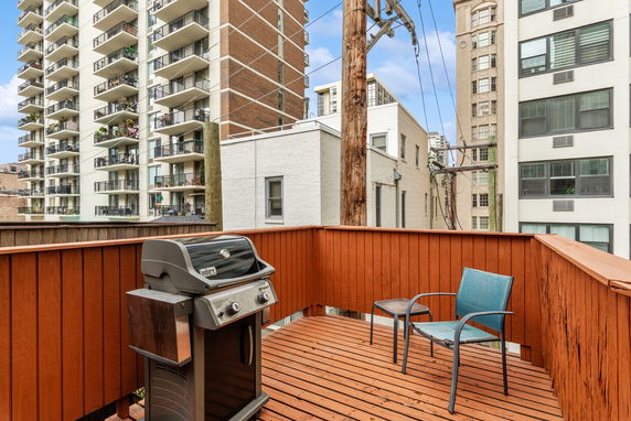 Rooftop deck with a barbecue grill and chair, surrounded by tall apartment buildings.