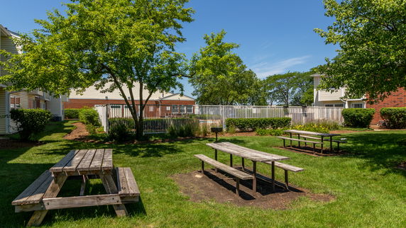 Picnic area with wooden tables and benches in a grassy outdoor space with nearby buildings.