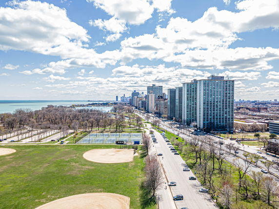 Wide view of a cityscape with tall buildings, a road, and a park area next to a body of water.
