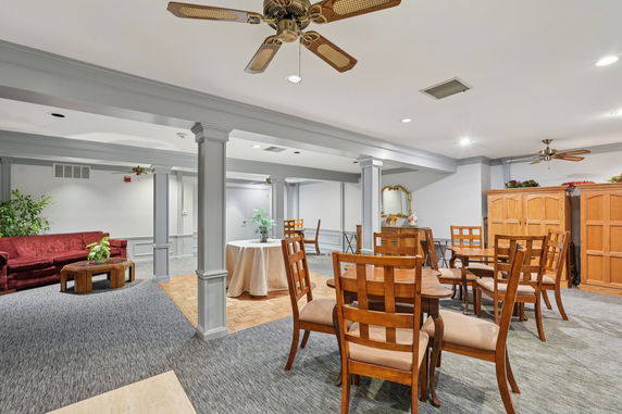 Interior view of a club house with tables and chairs, a couch, ceiling fans, and decorative plants.